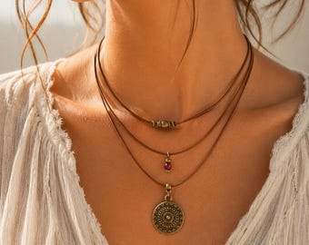 May include: A multi-strand necklace featuring three thin brown leather cords. The top cord has a small cylindrical brass bead, the middle cord holds a tiny red bead, and the bottom cord features a round, textured brass pendant with geometric patterns.