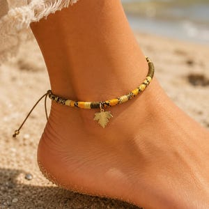 May include: An anklet featuring a braided cord in shades of yellow, olive green, and black, accented with a small gold-toned metal charm shaped like a maple leaf. The anklet includes adjustable thin cord ties at the back for a secure fit.