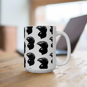 Può includere: Tazza in ceramica bianca con un motivo ripetuto di silhouette nere. Le silhouette raffigurano un profilo stilizzato con un'acconciatura a pompadour. La tazza ha un manico grande ed è posta su uno sfondo sfocato.