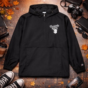 Può includere: Giacca a vento nera con cappuccio, chiusura a mezza zip e tasca a marsupio. La giacca presenta il testo bianco "Rockabilly Ramma" con una grafica di coniglio. La giacca è realizzata in materiale leggero e ha un logo Champion sulla manica.