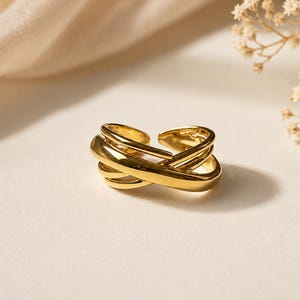 Puede incluir: Un anillo de tono dorado pulido que presenta un diseño cruzado de múltiples capas con varios aros lisos e interconectados que forman una estructura abierta y ajustable. La superficie metálica es reflectante y capta la luz, resaltando las líneas curvas.