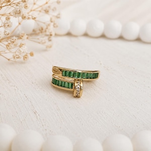 Puede incluir: Un anillo de oro tipo bypass con dos filas de piedras verdes en talla baguette engastadas en canal. Una pequeña barra vertical conecta los dos extremos, adornada con una fila de diminutas piedras redondas transparentes.