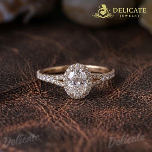 May include: A gold engagement ring featuring an oval-cut diamond centre stone, encircled by a halo of smaller diamonds. The band is also embellished with diamonds. The ring is set against a brown leather backdrop. The words "Delicate Jewelry" are visible.