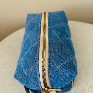 Könnte beinhalten: Eine gesteppte Kosmetiktasche aus blauem Denim mit goldenem Reißverschluss und beigem Stoff-Zuglasche. Die Tasche hat ein Rautenmuster und einen beigen Riemen. Die Tasche steht auf einer neutralfarbenen Oberfläche.