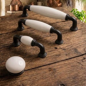 Puede incluir: Un conjunto de herrajes para armarios, que incluye tres tiradores y un pomo. Los tiradores presentan un centro blanco de forma ovalada y soportes negros curvados. El pomo es una esfera blanca con una base negra. Se muestra sobre una superficie de madera rústica.