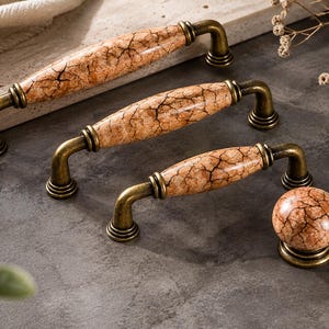 May include: Three cabinet pulls and one round cabinet knob featuring a smooth, light brown ceramic center with a dark brown crackle pattern. Each piece is finished with antique brass metal hardware, including curved handles and circular bases.