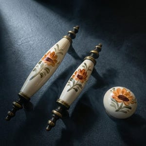 May include: Cream-coloured cabinet hardware set featuring a floral design with orange and brown flowers and green leaves. The set includes two handles and a round knob with brass-coloured accents.