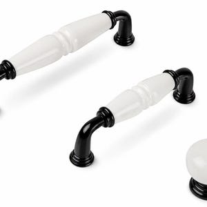 May include: Two cabinet pulls and one round cabinet knob. Each piece features a smooth white ceramic centre section with glossy black metal ends and circular mounting bases. The pulls have a curved, elongated shape, while the knob is spherical.