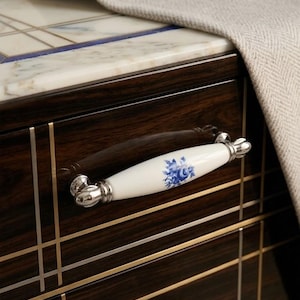 Puede incluir: Primer plano de un cajón de mueble de madera oscura con un tirador de porcelana blanca. El tirador presenta un diseño floral azul y detalles metálicos plateados. Un tejido gris claro descansa sobre la encimera de mármol encima del cajón.