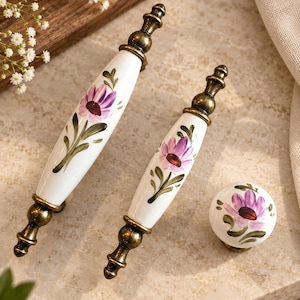 May include: Two elongated ceramic drawer pulls and one round ceramic knob, all featuring a white base with a hand-painted purple flower and green leaves. Each piece is accented with antique brass-toned metal hardware at the ends or base.