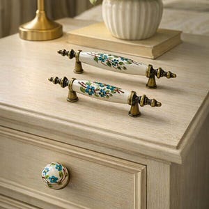 May include: A light-colored wooden nightstand with two floral-decorated drawer pulls and a matching knob. The drawer pulls are white with blue floral designs and brass-colored hardware. A lamp and a vase of flowers are on top.