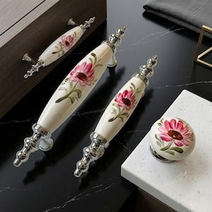 May include: Cream-coloured cabinet pulls and a knob with a floral design. The pulls have a silver base and are painted with pink flowers and green leaves. The knob has a matching design. The hardware is displayed on a black surface.