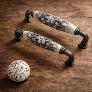 May include: Two cabinet handles and a knob with a floral design. The handles have a white and black floral pattern on a cream background, with dark gray metal hardware. The knob has a similar design.