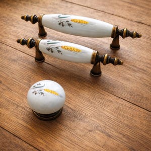 Könnte beinhalten: Weiße Keramik-Schrankgriffe und -Knopf mit Weizenmuster. Die Griffe haben eine bronzefarbene Basis und Enden. Das Weizenmuster ist gelb und grün. Der Knopf ist rund und hat das gleiche Weizenmuster. Die Beschläge befinden sich auf einer Holzoberfläche.