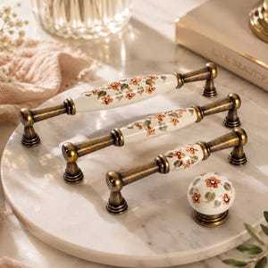 May include: Three cabinet pulls and one round cabinet knob featuring a white ceramic center with a hand-painted orange floral pattern and green leaves. The hardware includes antique brass-finished metal ends and mounting posts for attachment.