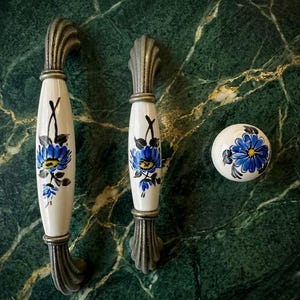 Puede incluir: Juego de tres piezas de herrajes decorativos para armarios. Incluye dos tiradores de cerámica blanca con diseños florales azules y un pomo redondo a juego. Los tiradores tienen bases y tapas de metal plateado ornamentado.