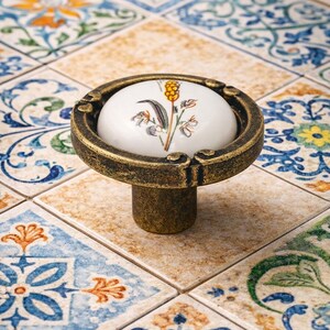 May include: A decorative cabinet knob with a white ceramic top featuring a botanical illustration. The knob has a bronze-colored metal base and is set against a backdrop of patterned tiles. The knob measures approximately 1.5 inches in height.