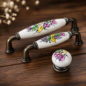 May include: Two white ceramic cabinet handles and a knob with a floral design in purple, yellow, and green. The handles have a curved, antique bronze base. The knob is round with a matching floral pattern.