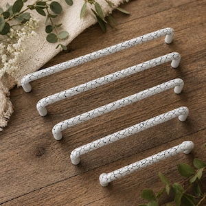 May include: A collection of white cabinet pulls with a distressed, crackle-like black pattern. The pulls are arranged diagonally on a wooden surface, showcasing their varying lengths. The hardware has a curved handle design.