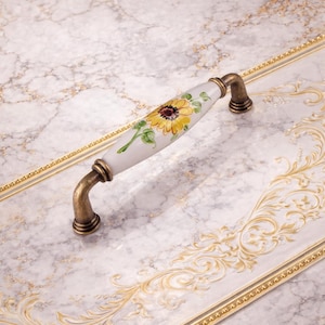 May include: A decorative cabinet pull with a white ceramic handle featuring a hand-painted sunflower design. The handle has brass-colored metal ends. The pull is set against a marble-patterned surface.
