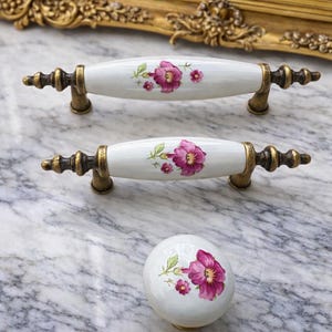 May include: White ceramic cabinet pulls and knob with a floral design in shades of pink and green. The hardware has antique brass-coloured metal accents. The pulls are oval-shaped, and the knob is round.