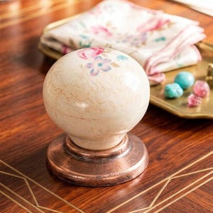 Puede incluir: Un pomo decorativo de color crema con un diseño floral en rosa y azul, sobre una base cobriza. El pomo se encuentra sobre una superficie de madera con incrustaciones doradas, con un paño con estampado floral y pequeñas baratijas en el fondo.