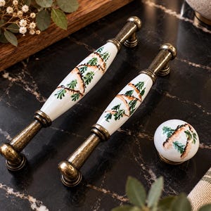 Può includere: Due maniglie per armadietto allungate e un pomello rotondo con centri in ceramica bianca, dipinti a mano con motivi di rami di pino verdi e dettagli di corteccia marrone, accentuati da finiture in metallo color ottone antico alle estremità.