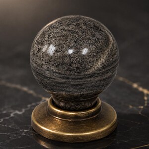 May include: A spherical cabinet knob featuring a polished grey and black mottled stone top with a smooth, reflective surface. The stone sphere is securely mounted onto a circular, brushed antique brass base with a warm, metallic finish.