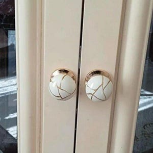Peut inclure: Deux boutons de placard ronds fixés sur des portes doubles de couleur crème clair. Chaque bouton présente une surface blanche lisse avec un motif craquelé doré et une base en métal doré poli fixée à la surface de la porte.
