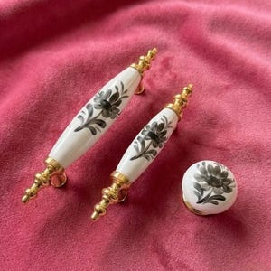 Puede incluir: Juego de tres piezas de herrajes decorativos para armarios. Incluye dos tiradores blancos y dorados con diseños florales negros y un pomo a juego. Los herrajes están sobre un fondo rosa.