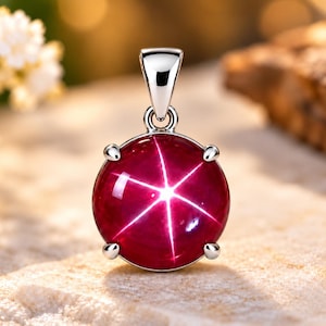 May include: A round, deep red star ruby pendant set in a polished silver-toned metal frame with four prongs. The gemstone features a distinct six-rayed star pattern that reflects light from the center, suspended by a silver-toned metal bail.