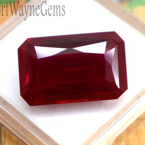 May include: A deep red, rectangular faceted gemstone with a brilliant cut, displayed inside a clear plastic storage box with a white foam insert. The text FortWayneGems is printed in black on the white foam surface of the display container.