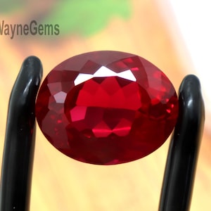 May include: A vibrant, oval-cut red gemstone with a faceted surface held by black tweezers. The stone displays deep crimson hues and light-reflecting facets. The text FortWayneGems is visible in the upper left corner of the frame.