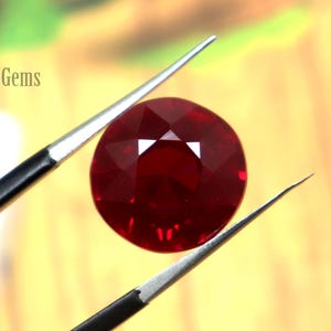 Puede incluir: Una gema facetada, redonda, de color rojo intenso es sostenida por unas pinzas. La gema es un rubí vibrante y pulido, que refleja la luz. El fondo es una mezcla borrosa de verde y marrón, con el texto "FortWayneGems" visible.