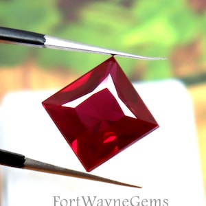 May include: A square-cut, deep red gemstone held by black metal tweezers. The gemstone is translucent with sharp, faceted edges. Below the gemstone is a white square container with the text FortWayneGems printed on the front surface.
