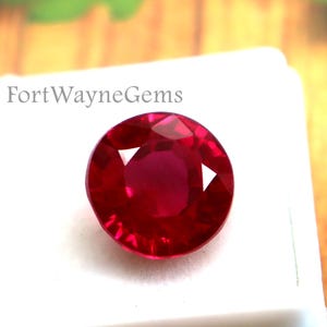 May include: A round, faceted, deep red gemstone sits inside a white plastic display box. The text FortWayneGems is printed in black on the white surface of the box. The gemstone displays a brilliant, reflective surface with multiple facets.
