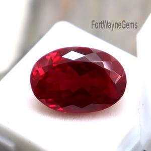 Puede incluir: Una gema roja oscura, facetada y de forma ovalada. La gema se muestra en una caja de plástico blanco con tapa transparente. El texto "FortWayneGems" es visible en la caja. La gema se encuentra sobre un fondo neutro.