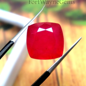 May include: A vibrant red, cushion-cut gemstone held by a pair of black-handled metal tweezers. The gemstone features a faceted surface that reflects light. The text FortWayneGems is visible in the background above the gemstone.