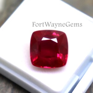 May include: A cushion-cut vibrant red gemstone with a faceted surface, displayed inside a small white square plastic display case. The text FortWayneGems is printed in black on the white insert of the case, which holds the loose gemstone securely.