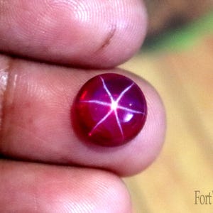 May include: A round, deep red star ruby gemstone featuring a distinct six-rayed asterism effect that creates a bright white star pattern across the center of the polished, cabochon-cut stone. The text FortWayneGems is visible in the corner.