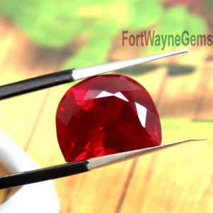 Peut inclure: Une pierre précieuse rouge vif, taillée en coussin, tenue par une pince en métal noir. La pierre présente une surface à facettes qui reflète la lumière, et le texte FortWayneGems est visible dans la partie supérieure droite de l'image.