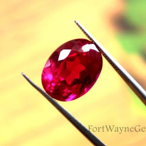 May include: A faceted, oval-shaped ruby gemstone held by tweezers. The ruby is a vibrant red color with a polished surface. The background is a blurred mix of yellow and green.