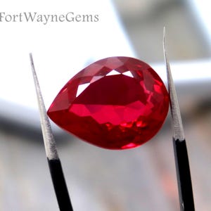 Puede incluir: Una piedra preciosa roja facetada en forma de pera sostenida por pinzas de metal con puntas negras. La piedra es translúcida y de color rojo vibrante. Al fondo, una caja de presentación cuadrada blanca muestra el texto FortWayneGems.