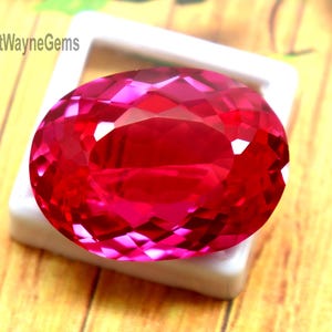 Puede incluir: Una gema roja vibrante, de corte ovalado y facetada, exhibida en un soporte cuadrado de plástico blanco. La gema muestra un tono carmesí profundo y brillante con facetas geométricas complejas. El texto FortWayneGems es visible.