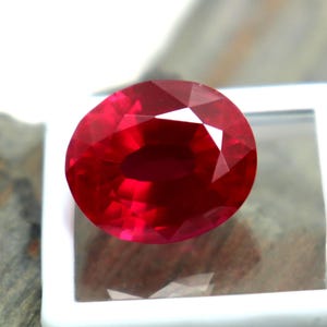 May include: A vibrant, oval-cut red gemstone with a faceted surface that reflects light. The gemstone is displayed resting on a small, square white plastic container with a clear top, showcasing its deep crimson color and polished clarity.