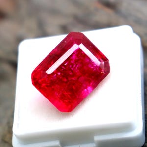 May include: A vibrant, rectangular-cut red gemstone with a faceted surface, displaying deep crimson hues and internal light reflections. The stone is positioned on a small, square white display base with a matching white lid resting nearby.