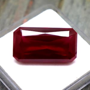 May include: A rectangular, faceted, deep red gemstone with a polished surface. The stone is displayed on a white square insert inside a clear, square plastic display case, showcasing its vibrant colour and precise geometric cut.