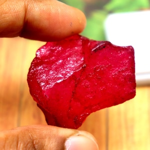 May include: A raw, uncut ruby gemstone with a deep, vibrant red color and an irregular, multifaceted surface texture. The stone displays natural inclusions and a translucent quality, showcasing a jagged, organic shape with sharp edges and corners.