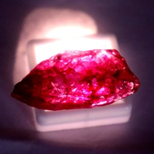 May include: A raw, unpolished gemstone with a vibrant, deep pinkish-red hue. The stone features a jagged, crystalline texture with translucent facets that catch the light, resting atop a small, rectangular white display base.