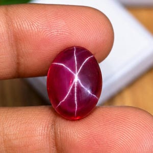 Pode incluir: Uma pedra preciosa de rubi estrela de formato oval em vermelho profundo, apresentando um efeito de asterismo de seis raios em sua superfície de cabochão lisa e polida. A gema exibe um tom carmesim saturado com um padrão de estrela branco.